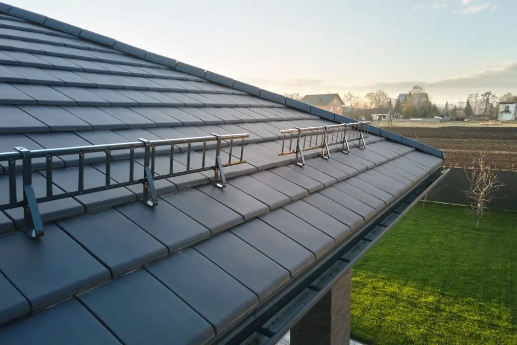 snow guards for metal roof A metal snow guard is installed along the edge of a dark tiled sloped roof on a house, overlooking a green lawn and open fields under a clear sky.