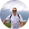 A person wearing sunglasses and a white t-shirt stands outdoors in a mountainous area. They are smiling, with a backpack on their shoulders. Clouds and a hazy mountain range are visible in the background.