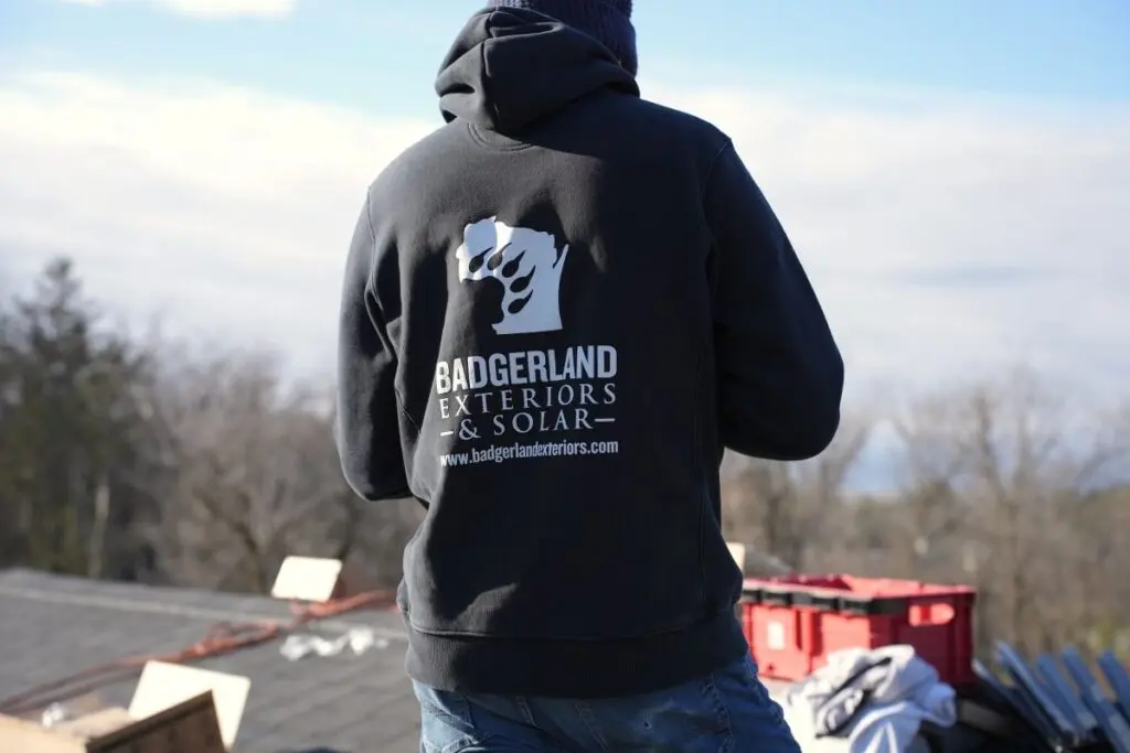 roof replacement timeline person wearing a black hoodie with the "Badgerland Exteriors & Solar" logo and website stands outdoors near construction materials, facing away from the camera. Trees and cloudy sky are in the background.