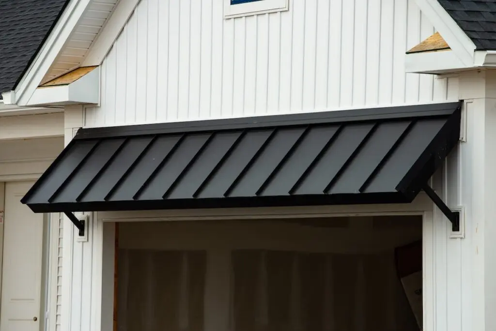 A black metal awning is installed above the entrance of a white building with vertical siding. The area beneath the awning appears to be an unfinished garage or storage space.
