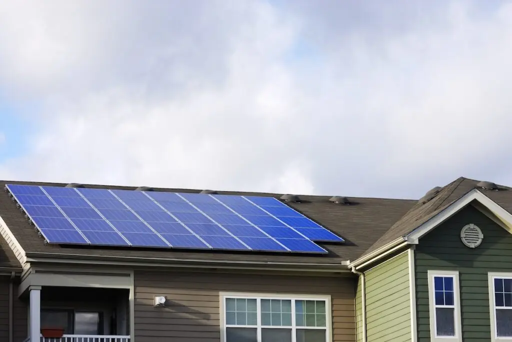 A rooftop featuring a solar installation in Stoughton showcases blue panels on a two-story house. Painted in gray and green, with crisp white window frames, the house stands under a partly cloudy sky.