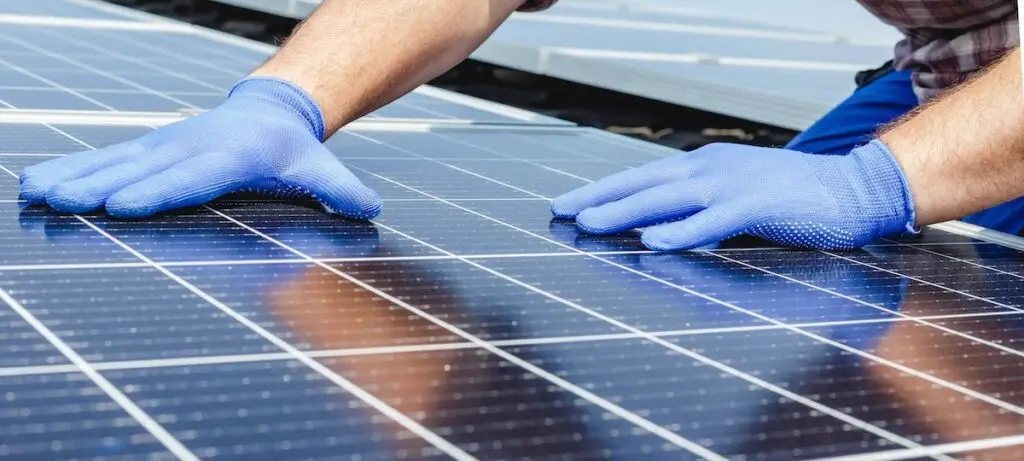 A person wearing blue gloves is inspecting large solar panels on a commercial roofing project in Madison. Their hands are placed on the panels, which reflect sunlight.