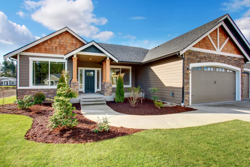 A charming suburban house with a stone and shingle exterior, a two-car garage, and a well-maintained front yard. The yard features neatly trimmed bushes and a freshly mulched garden bed. A curved walkway leads to the front porch.