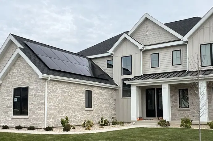 Modern two-story house with light-colored brick and siding, featuring solar panels on the roof above the garage. The front yard has neatly trimmed grass, small shrubs, and a covered entryway.