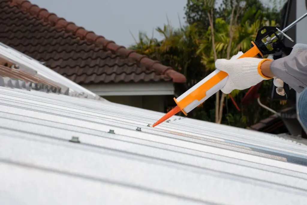 waterproof sealant for metal roof A person wearing white gloves applies waterproof sealant for metal roof to the joints of a white metal roof using a caulking gun. There are trees and a house with a brown tiled roof in the background.