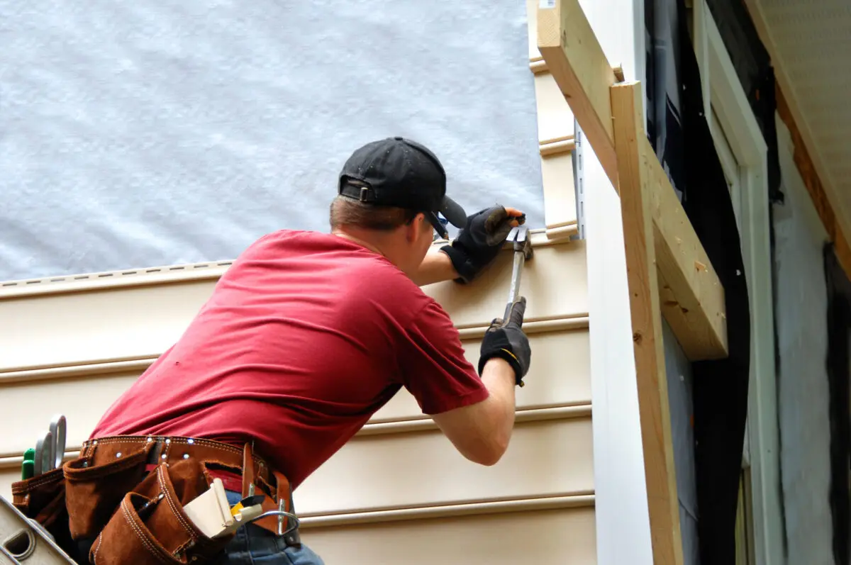 A construction worker in a red shirt and tool belt uses a hammer to install beige vinyl siding on a house exterior. He is facing the wall and wearing a black cap.