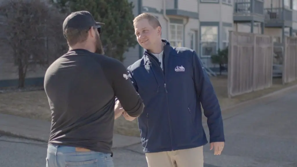 do roofers need to be licensed in sun prairie wi Two men shake hands outdoors near a residential building. One wears a navy jacket and khakis, while the other wears a black shirt, jeans, and a black cap. They appear to be greeting each other on a sunny day.