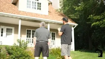 Two people stand outside a house with a beige exterior and a sloped, shingle roof. They face the house, framed by trees and shrubs, under a clear sky. Both are wearing casual outfits with shorts and dark shirts.