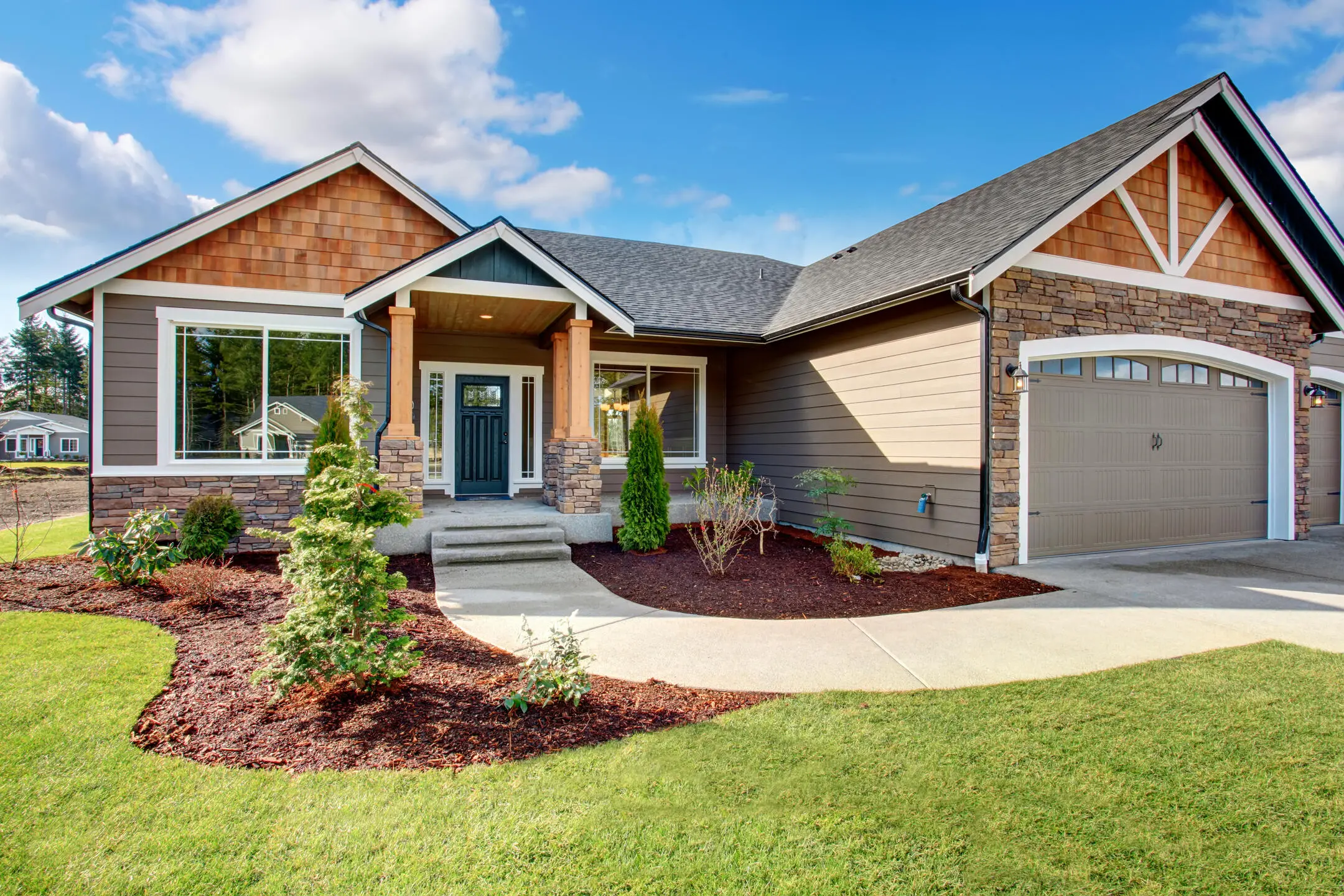 A charming suburban house with a stone and shingle exterior, a two-car garage, and a well-maintained front yard. The yard features neatly trimmed bushes and a freshly mulched garden bed. A curved walkway leads to the front porch.