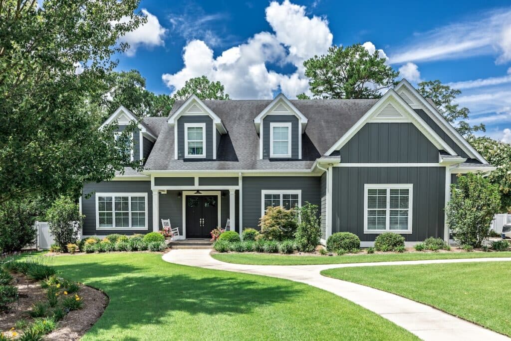 A gray two-story suburban house with white trim, front porch, dormer windows, and neatly landscaped lawn under a blue sky with clouds.