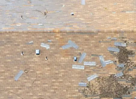 Aerial view of a shingle roof with several missing and damaged shingles, exposing the underlayment beneath. Scattered debris and a few vents are visible across the roof.