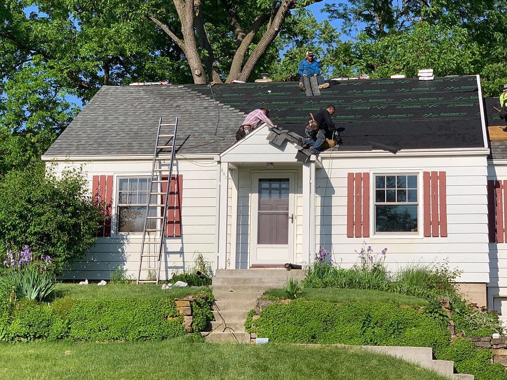 Three workers are installing new roofing material on a small white house with red shutters, surrounded by greenery and colorful flowers, with ladders propped against the roof.