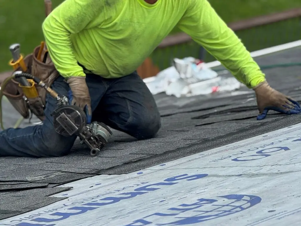 roof replacement timeline construction worker in a bright yellow shirt installs asphalt shingles on a roof, holding a nail gun and surrounded by roofing materials and tools.