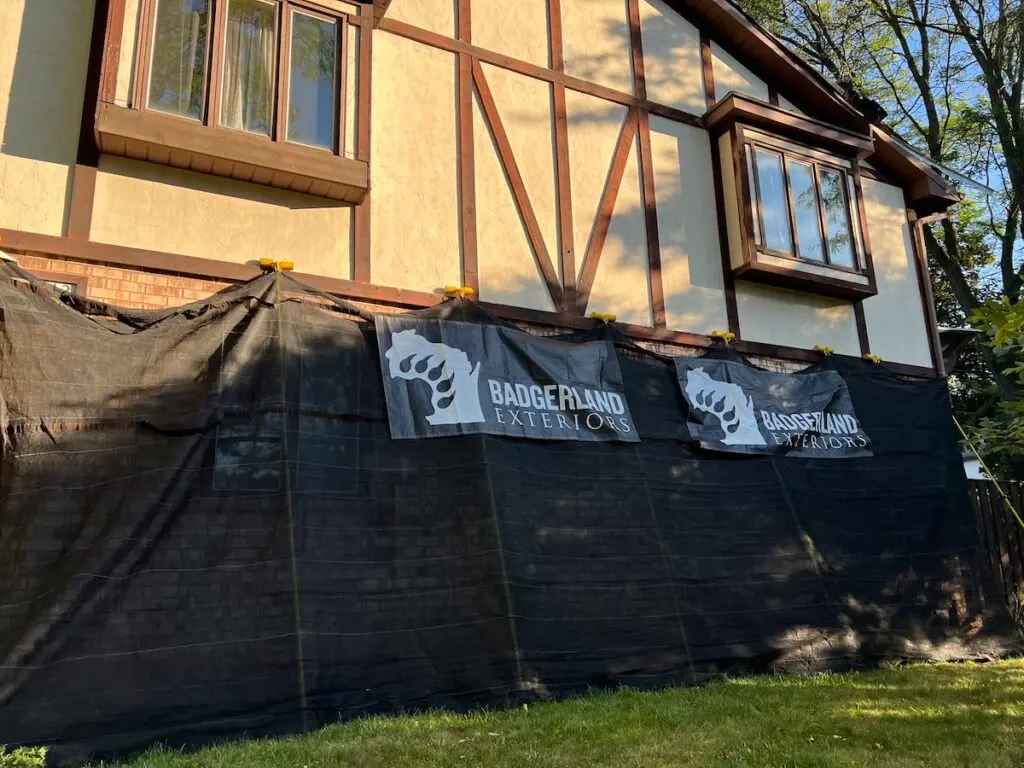 A house with Tudor-style architecture is partially covered by black construction fencing. Two "Badgerland Exteriors" banners hang on the fence. Sunlight and tree shadows are visible on the building’s facade.