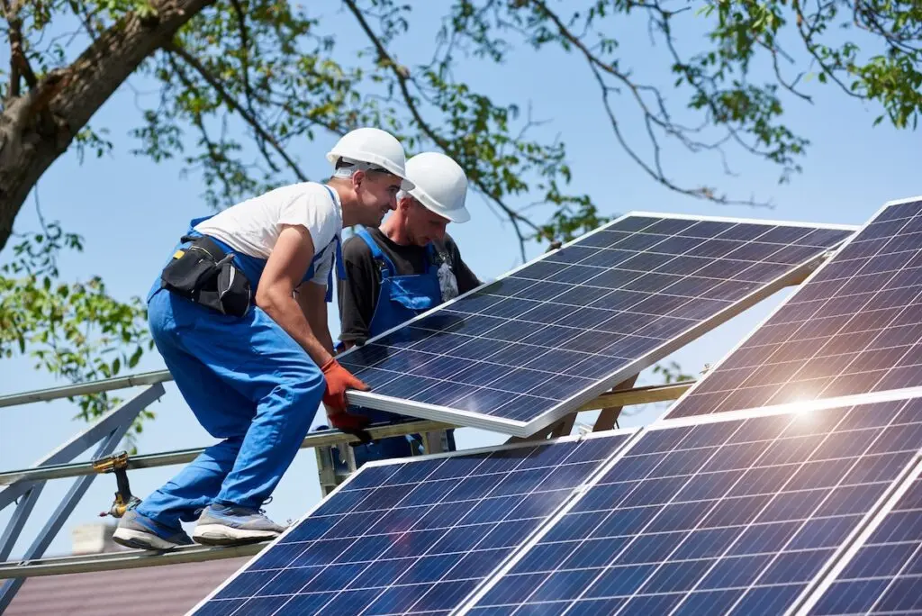 Two workers in blue overalls and white helmets are expertly installing solar panels on a commercial roofing site in Madison. Under a clear sky, they adjust the panels on a metal frame, with green tree branches as their backdrop. Sunlight reflects brilliantly off the panels.