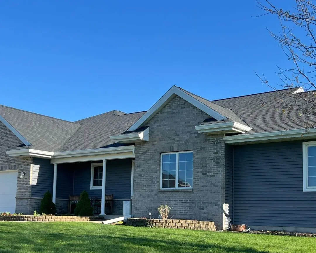 A modern single-story house with gray brick and blue siding, a dark roof—expertly done by roofing New Berlin—white trim, and a small front porch with two chairs. The yard is green and well-kept under a clear blue sky.