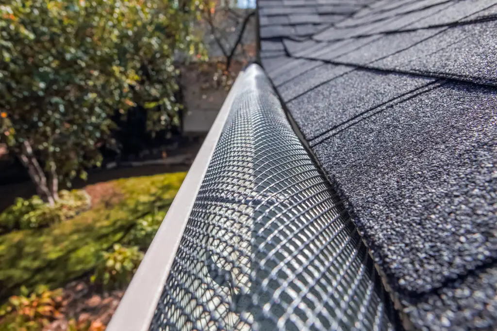 Close-up view of a mesh gutter guard installed along the edge of a shingled roof, hinting at how to install gutter guards, with a blurred garden and trees visible in the background. Plastic guard over gutter on a roof with a leaf stuck on the out