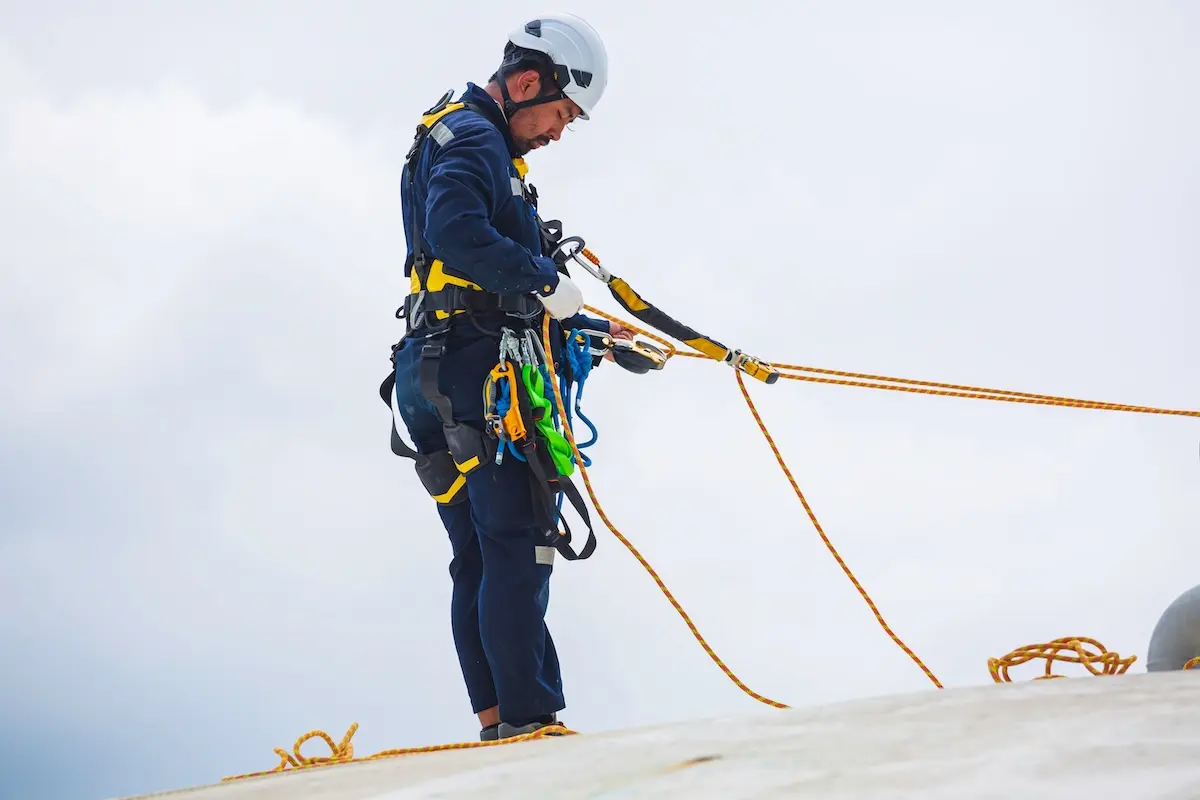 A worker wearing safety gear and a helmet is using ropes and harnesses while standing on a sloped surface, performing maintenance or inspection work at height against a cloudy sky.