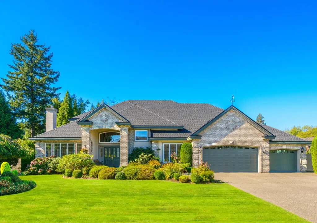 A large, modern brick house with a dark gray roof, three-car garage, and well-maintained lawn. Surrounded by neatly trimmed bushes and trees, the driveway leads to the front entrance under a clear blue sky.