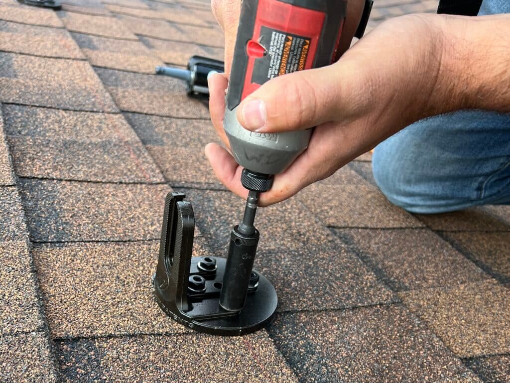 roof maintenance checklist A person uses a power drill to install a black metal bracket onto a brown shingled roof, with one hand holding the drill and the other steadying the bracket.