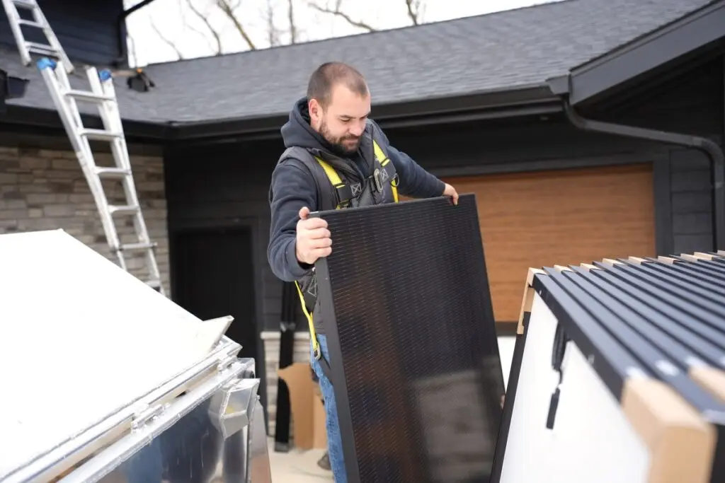A man wearing a safety harness lifts a solar panel near a house with a dark roof, a ladder, and other solar panels stacked nearby.