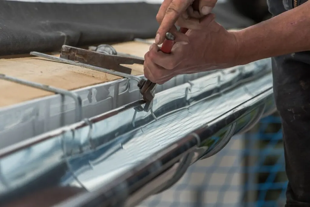 gutter replacement Close-up of a person’s hands using pliers to install or repair a shiny metal rain gutter on a building, with a wooden board and protective covering visible nearby.