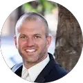 A man in a suit and tie is smiling with a blurred outdoor background featuring a tree and some greenery.