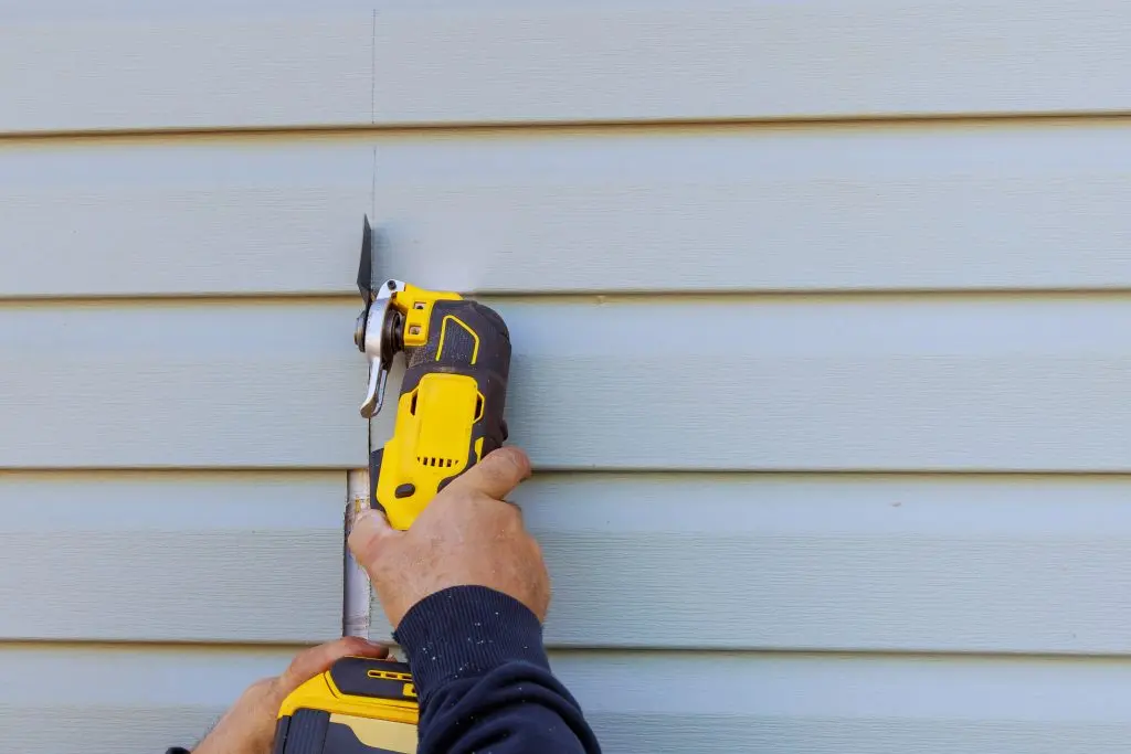 vinyl sidint cost per square foot A person uses a yellow power tool to cut into blue vinyl siding on a house exterior.
