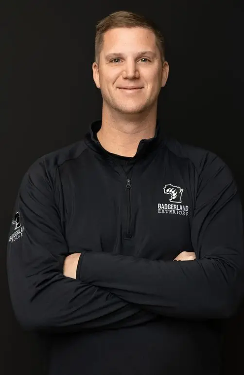 A man with short light brown hair stands confidently with arms crossed, wearing a black zip-up jacket with 