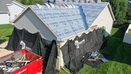 A house with its roof under construction, covered in underlayment and roofing materials. Black tarps protect the sides, debris is scattered below, and a red dumpster holds old roofing material.