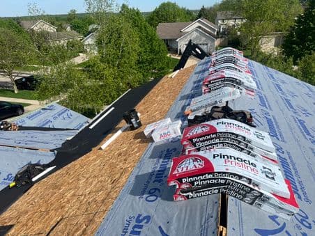 roofing companies sun prairie wi Roof under construction with stacks of shingles, tools, and materials placed on a partially completed roof. Surrounding trees and houses are visible in the background under a clear sky.