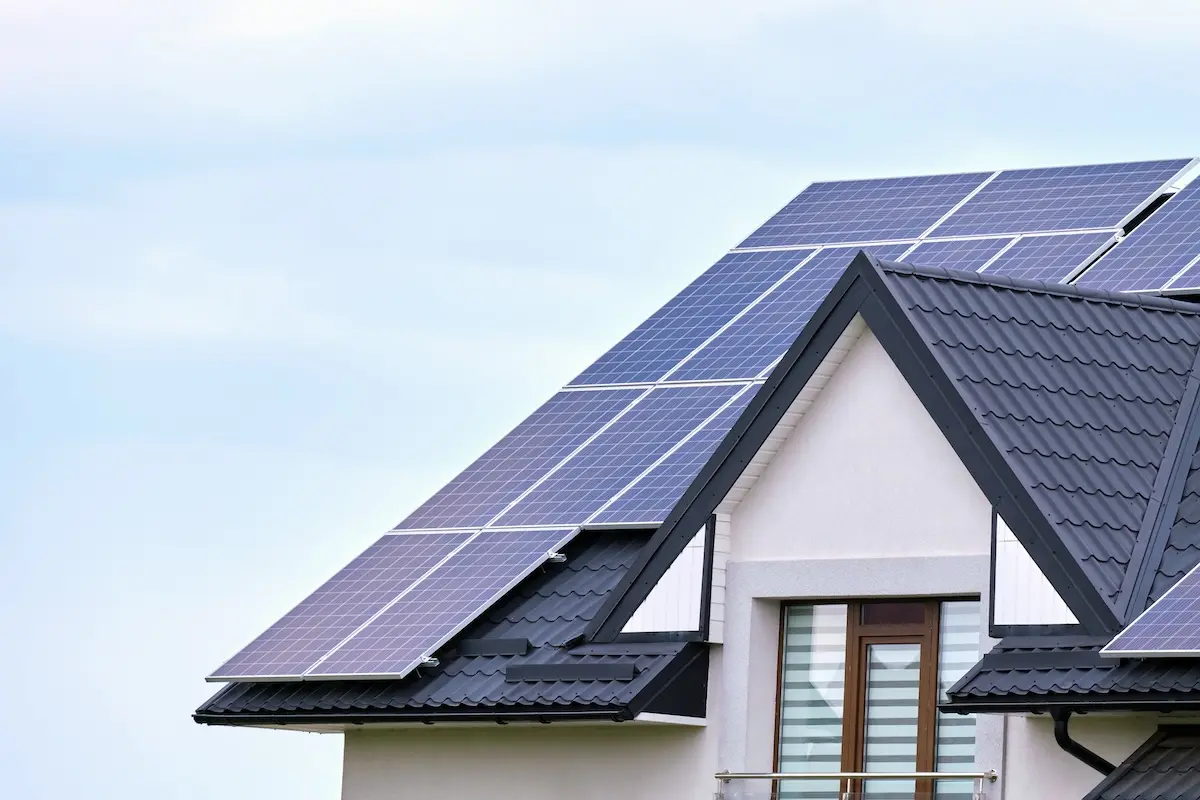 A modern house with a dark gray roof featuring several solar panels arranged on top. Beneath the panels, there's a window with partially drawn blinds. The sky is clear and blue in the background.