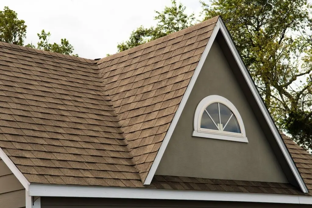 A close-up view of a house roof with best roof shingles in brown, featuring a gabled design and an arched window with white trim set against a grayish exterior wall. Trees and sky are visible in the background.