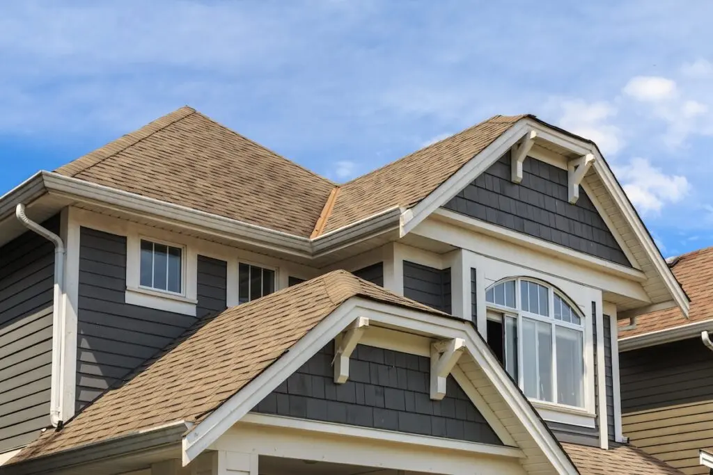 siding types Close-up of a modern house exterior featuring expert siding installation with dark gray siding, white trim, multiple gables, and a brown shingled roof against a blue sky with scattered clouds.
