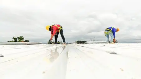commercial roof repair services Two construction workers wearing safety gear and harnesses are working on a white metal roof under a cloudy sky. One is crouched, using tools, while the other is bent over, both focused on their tasks.