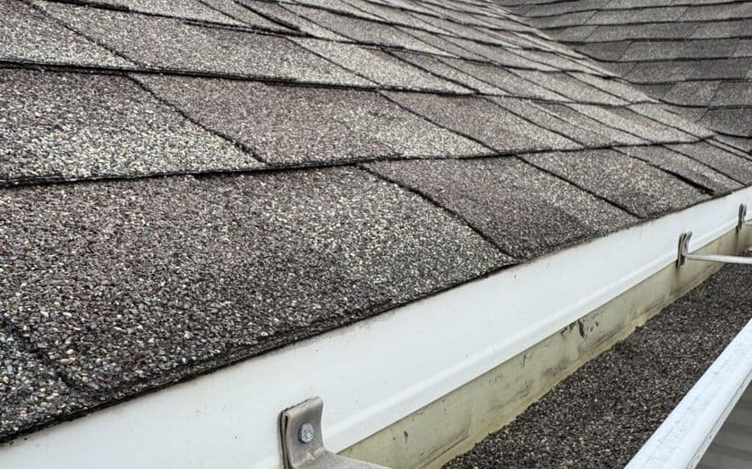 Close-up view of a shingled roof with an attached white gutter, containing leaves and debris. The roof shingles appear worn and textured, and the gutter shows some discoloration and buildup.