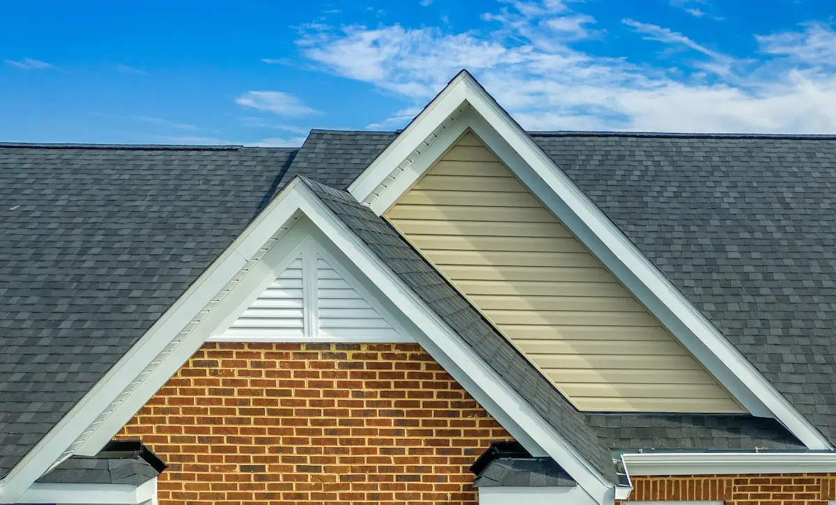 siding types Close-up of a house’s roof with overlapping gable peaks, highlighting expert siding installation with beige siding, dark shingles, red brick walls, and white trim, all set against a blue sky with scattered clouds.