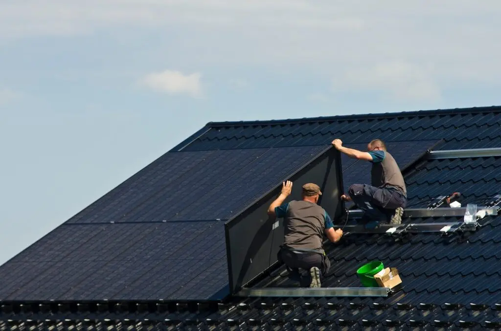 Two workers in uniform conduct a solar installation in Fithburg, mounting panels on a sloped, dark-colored roof under a clear sky. Various tools and a green bucket are scattered nearby, enhancing the glossy finish of the multiple panels on the roof.