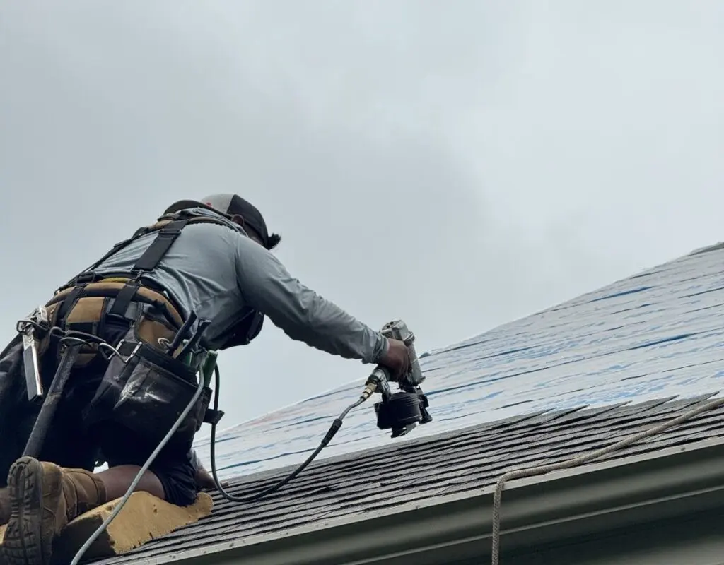 A person wearing a safety harness and tool belt is kneeling on a roof, using a nail gun to install shingles under a cloudy sky—demonstrating expert roofing Madison residents can rely on.