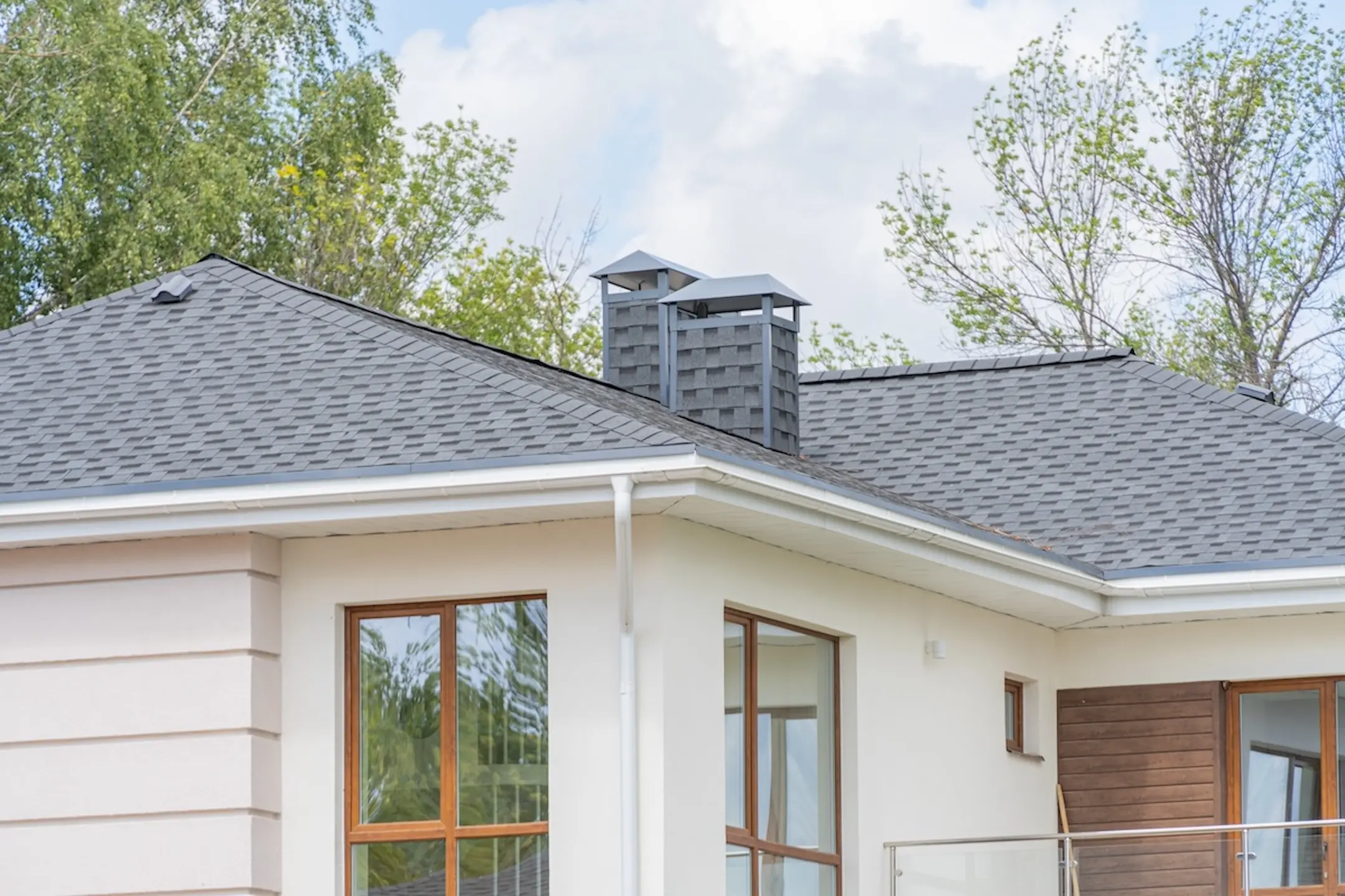A modern house with gray shingle roofing Oconomowoc style, two metal chimney caps, large windows, and light-colored walls, surrounded by trees under a partly cloudy sky.