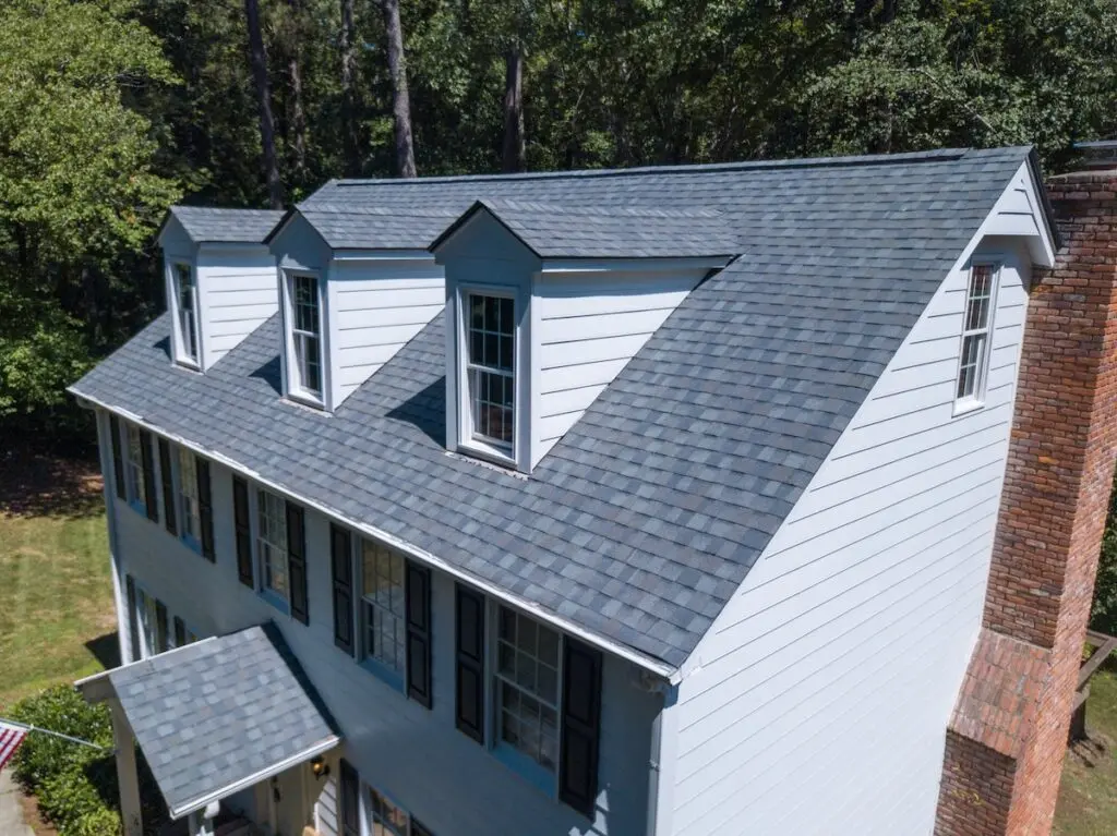 Aerial view of a white house with best roof shingles, grey shingle roof, three dormer windows, black shutters, brick chimney, and surrounded by green trees.