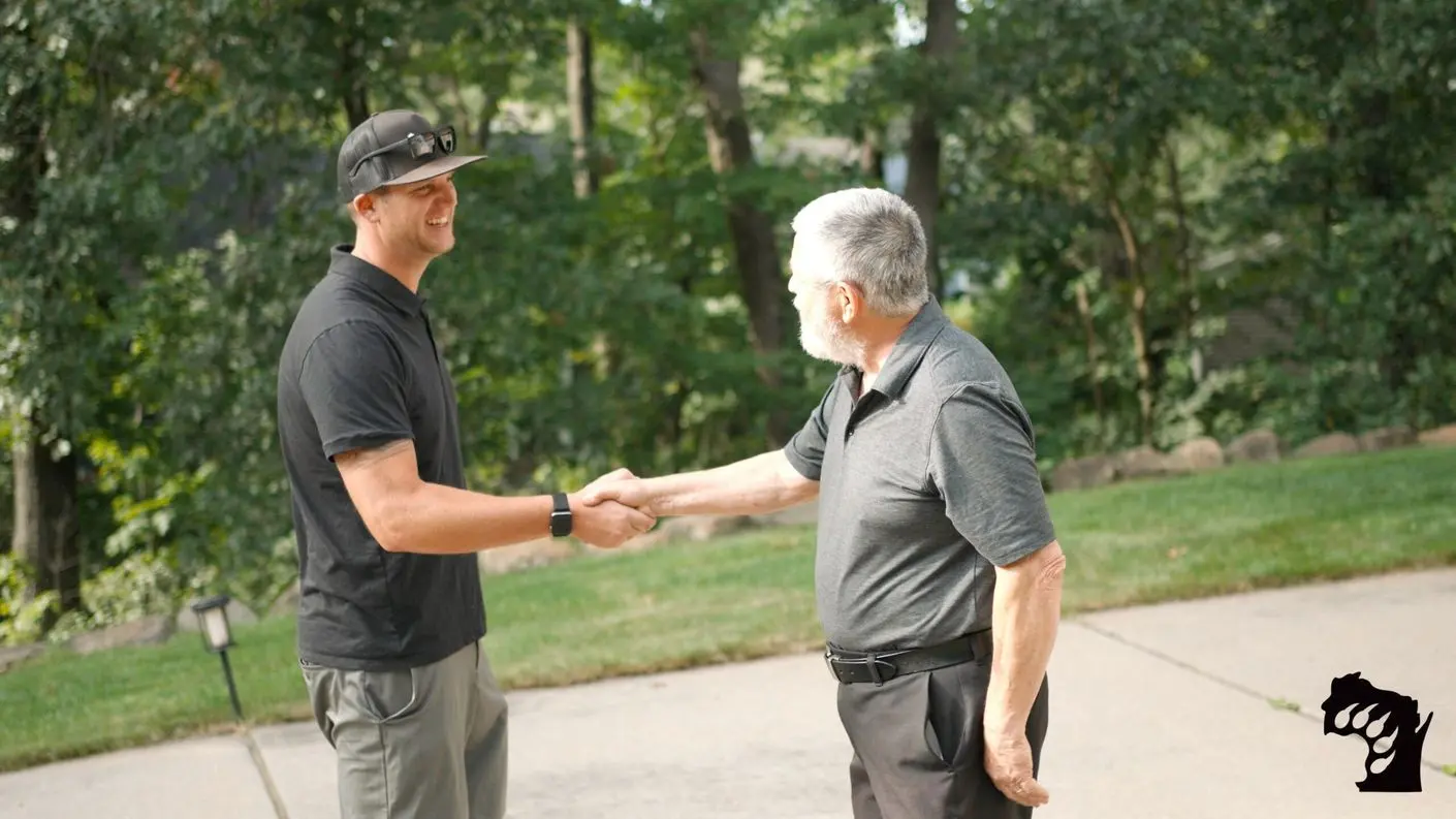 Two men wearing black shirts shake hands outdoors on a sunny day. One man wears a cap and sunglasses, while the other has gray hair and a beard. They are standing on a paved path with trees and grass in the background.