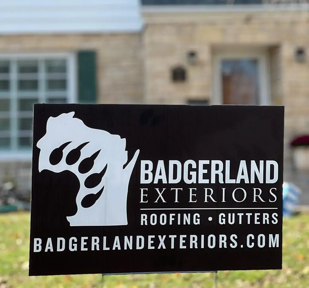 A sign reads "Badgerland Exteriors - Roofing • Gutters" with their website listed below. The sign features a silhouette of Wisconsin and is placed in front of a brick building.