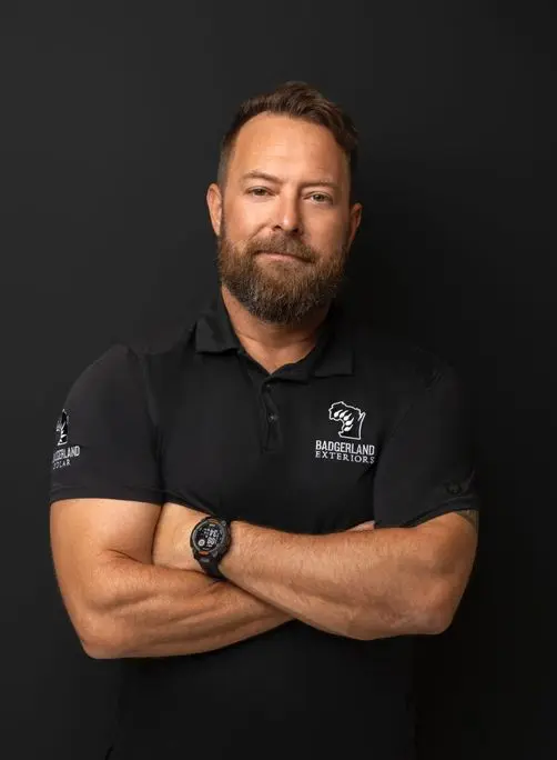 A bearded man with short hair stands against a black background, arms crossed, wearing a black polo shirt with 