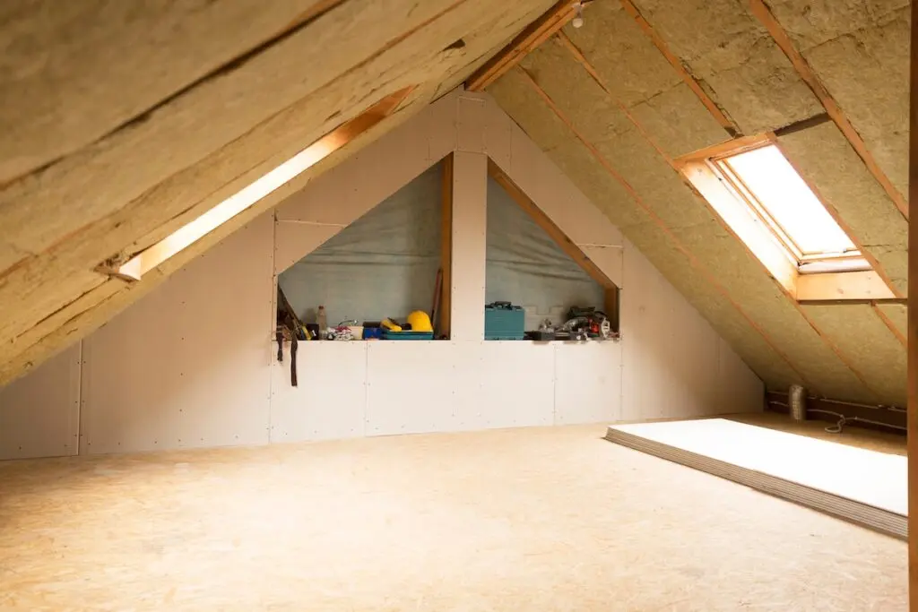 An unfinished attic with slanted insulation, a skylight, partially installed drywall, tools and materials on a shelf, and some stacked boards on the floor.