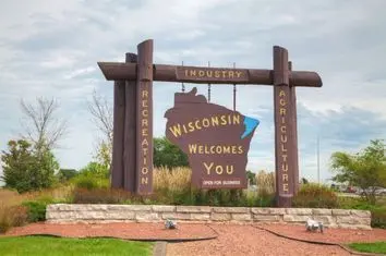 A tourist sign in a landscaped area welcomes visitors to Wisconsin. It features a wooden map of the state with the words "Wisconsin Welcomes You," highlighting industry, agriculture, and recreation. A stone base and greenery surround the sign.