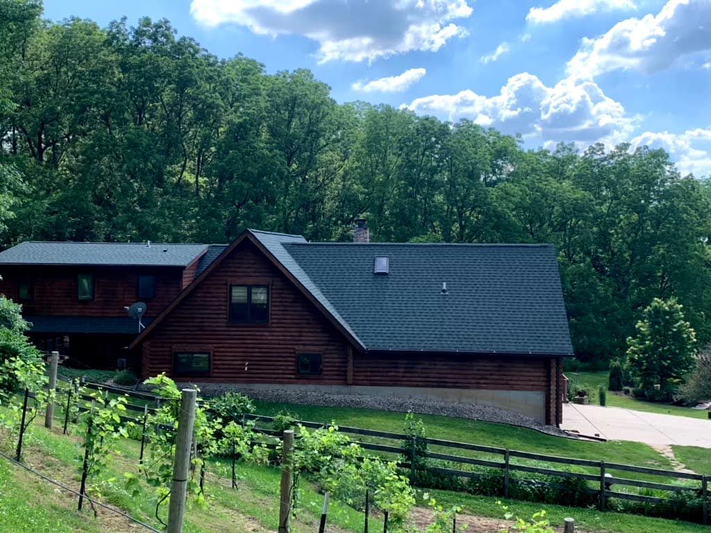 A large wooden cabin with a dark roof, installed by roofing Waukesha experts, sits nestled among green trees under a partly cloudy sky, with a sloped yard, wooden fence, and small plants in the foreground.