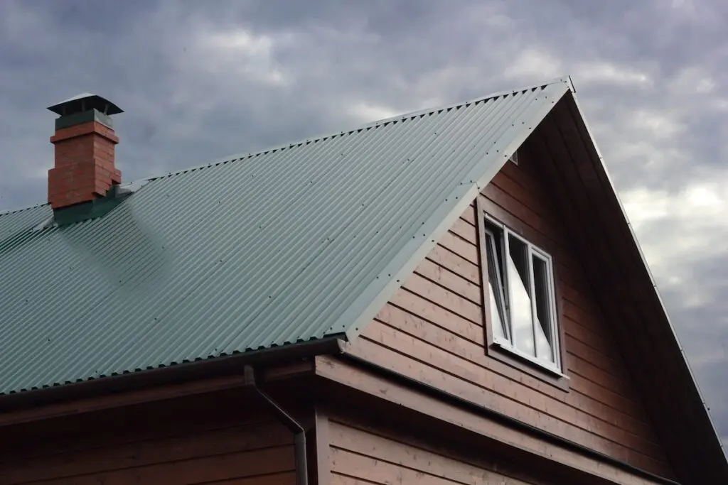 A close-up view of a wooden house with a green corrugated metal roof, showcasing one of the popular types of metal roofing, a brick chimney, and a double window set against a cloudy sky.
