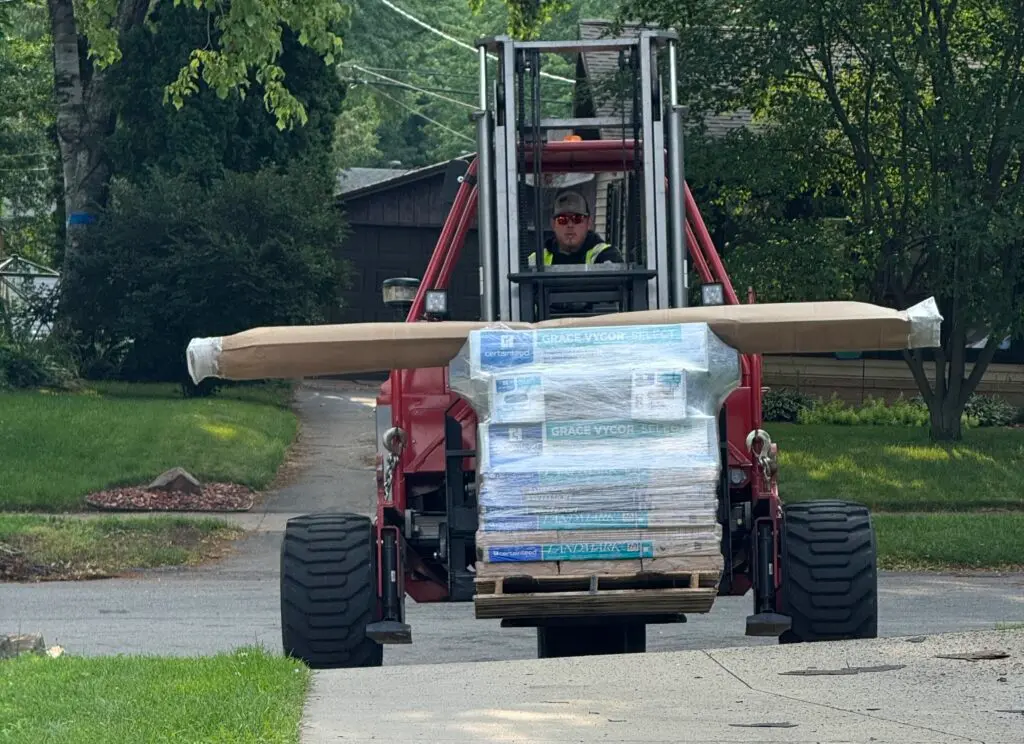 A worker drives a red forklift carrying a large pallet of construction materials, including long wrapped boards, down a residential driveway lined with green grass and trees.