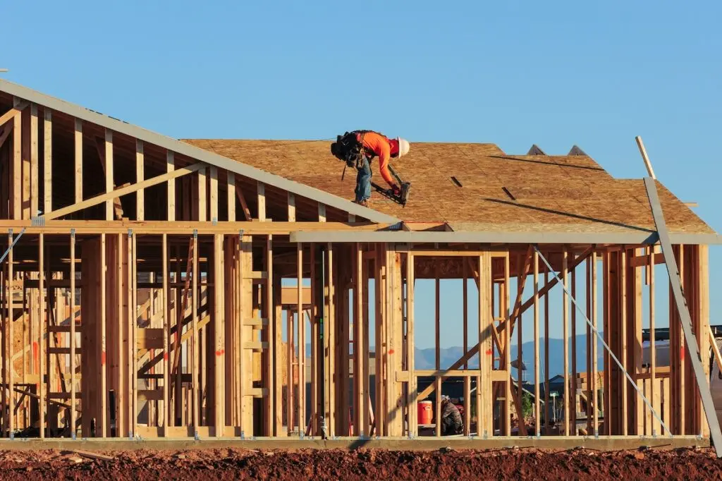 A construction worker in a hard hat is kneeling and working on the roof of a wooden house frame under clear blue skies. The structure is incomplete, showing exposed beams and framework.